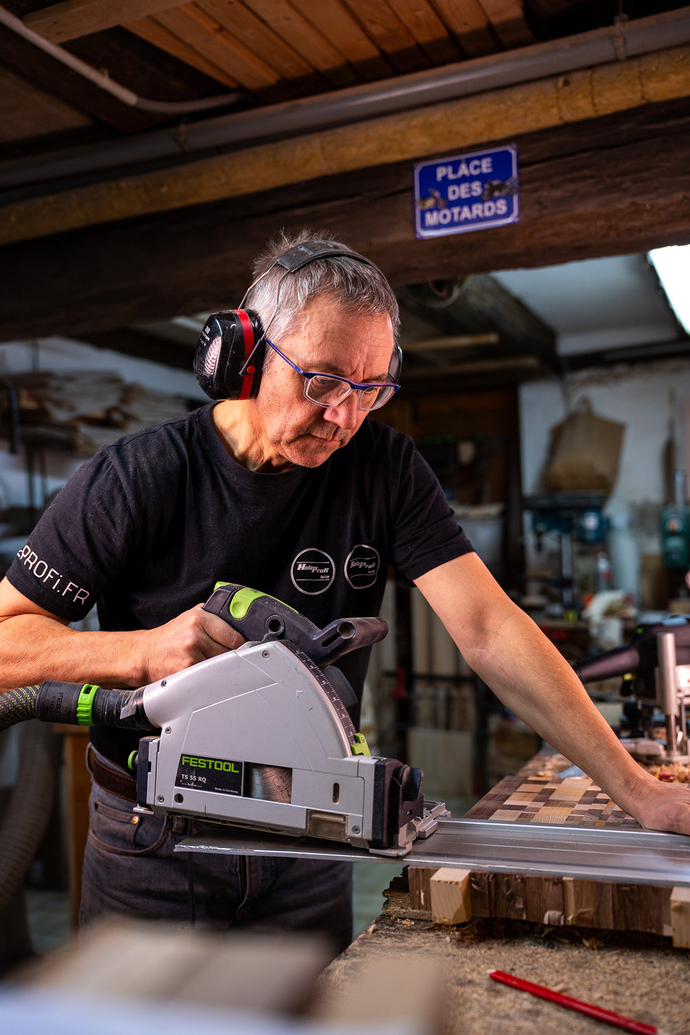A man wearing glasses and hearing protection uses a Festool power saw to cut a wooden board in a workshop.
