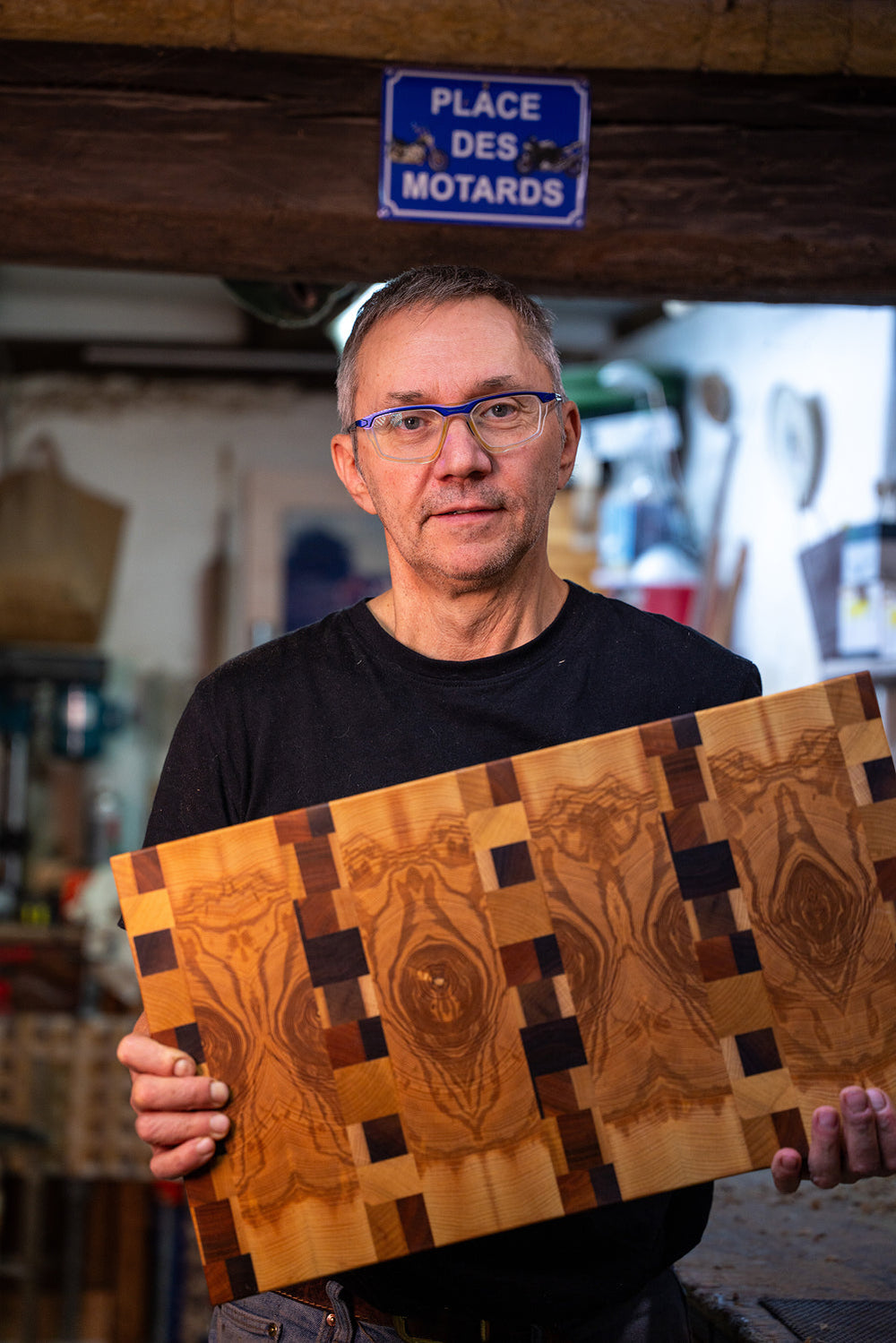 Man wearing glasses holding an intricately patterned wooden board in a workshop.